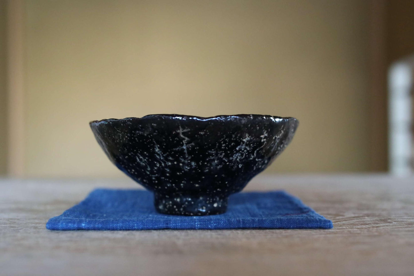 A black bowl with white speckles sits on a blue cloth on a wooden surface.