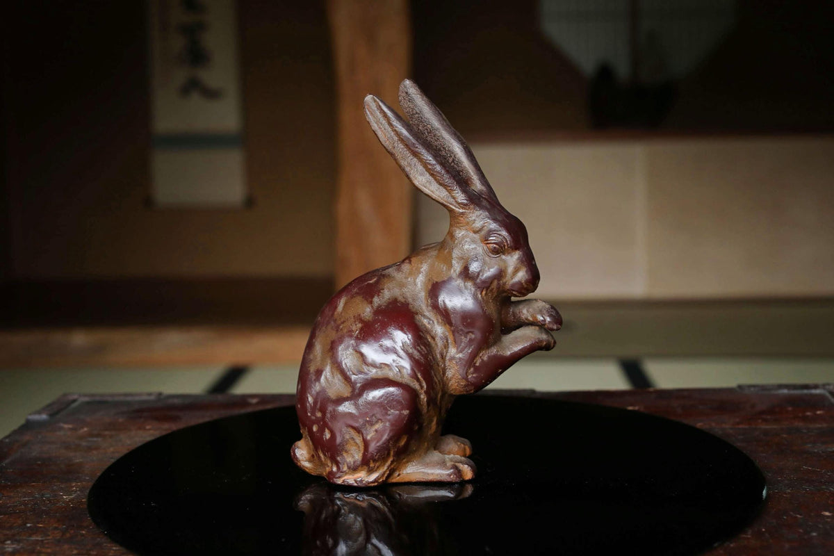 A ceramic rabbit figurine is sitting on a black circular surface, with a blurred background that appears to be a room with a wooden floor and a wall with a window.