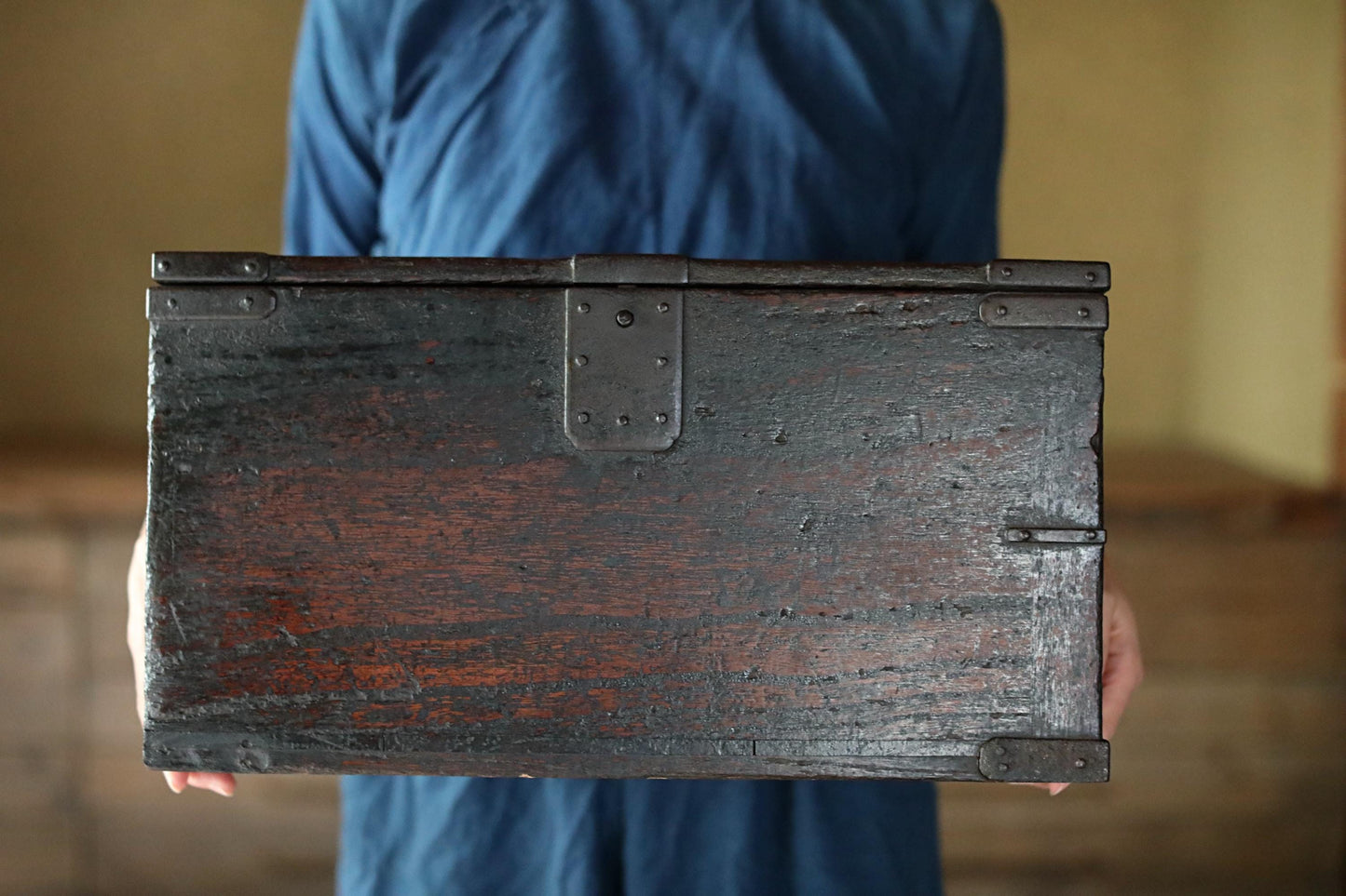 A person wearing a blue shirt is holding a large, old-fashioned wooden chest with metal clasps.