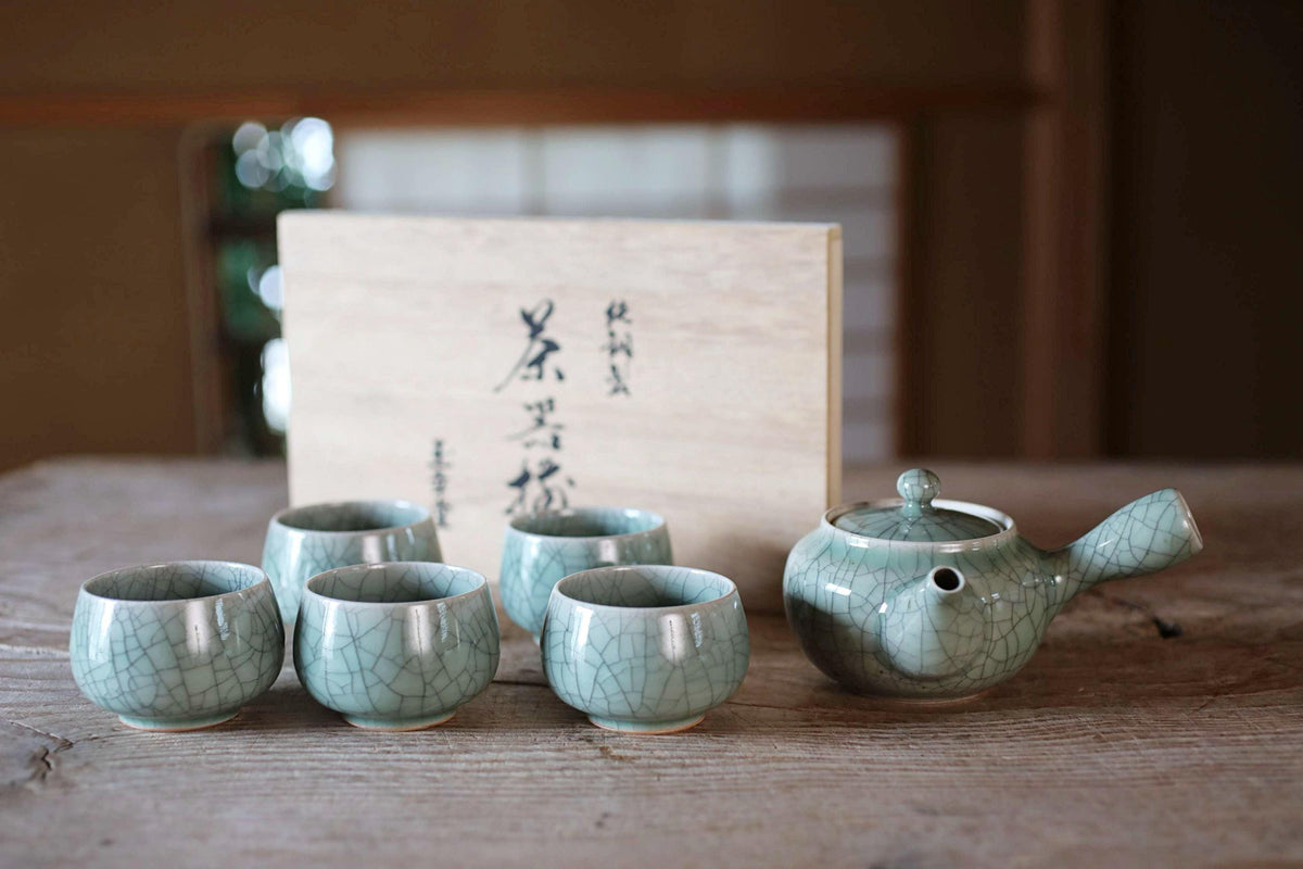 A set of six green ceramic cups arranged on a wooden table, with a teapot and a wooden box with Japanese characters in the background.