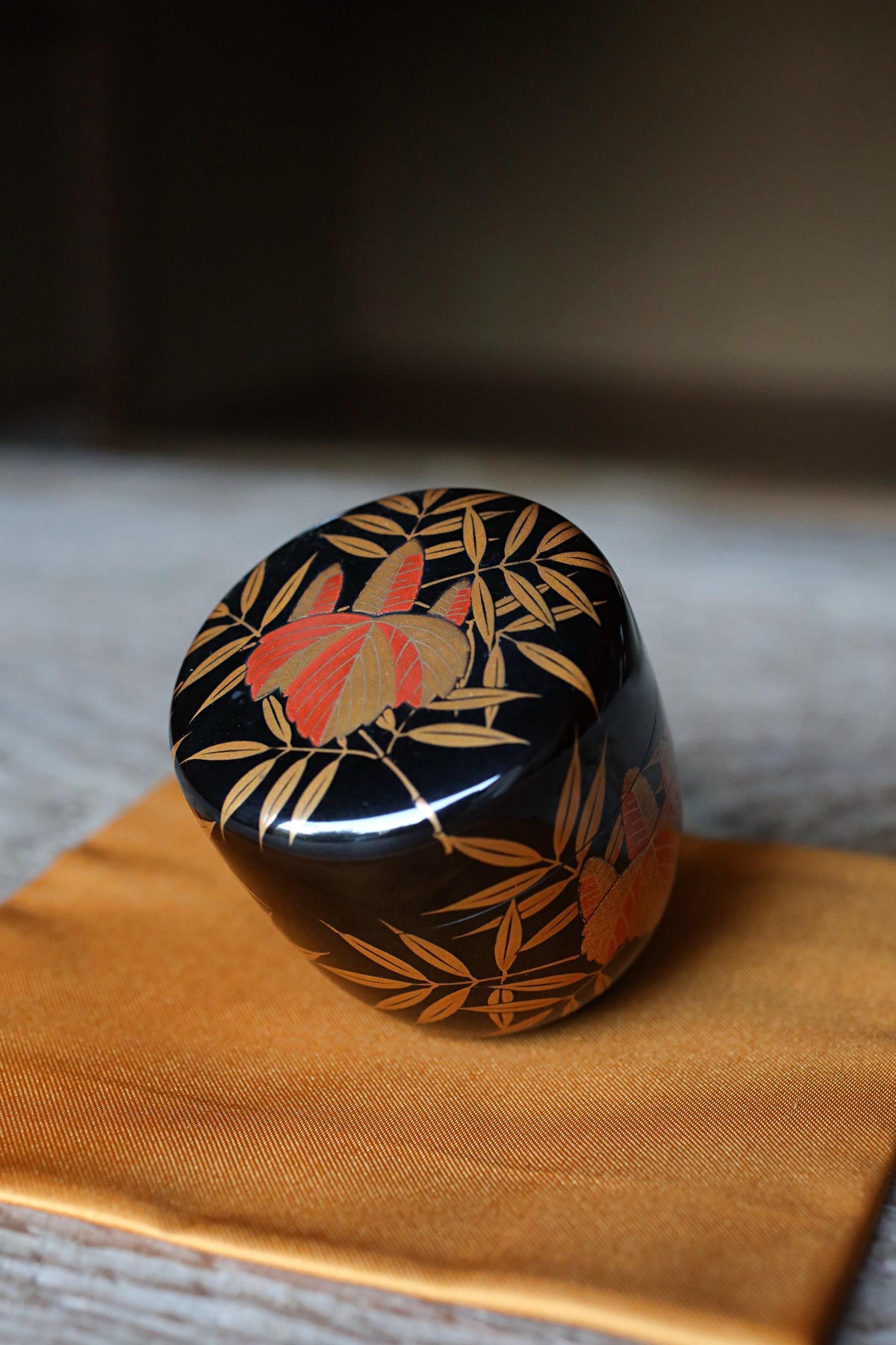 A decorative black and gold ceramic container with a floral pattern sits on a yellow cloth.