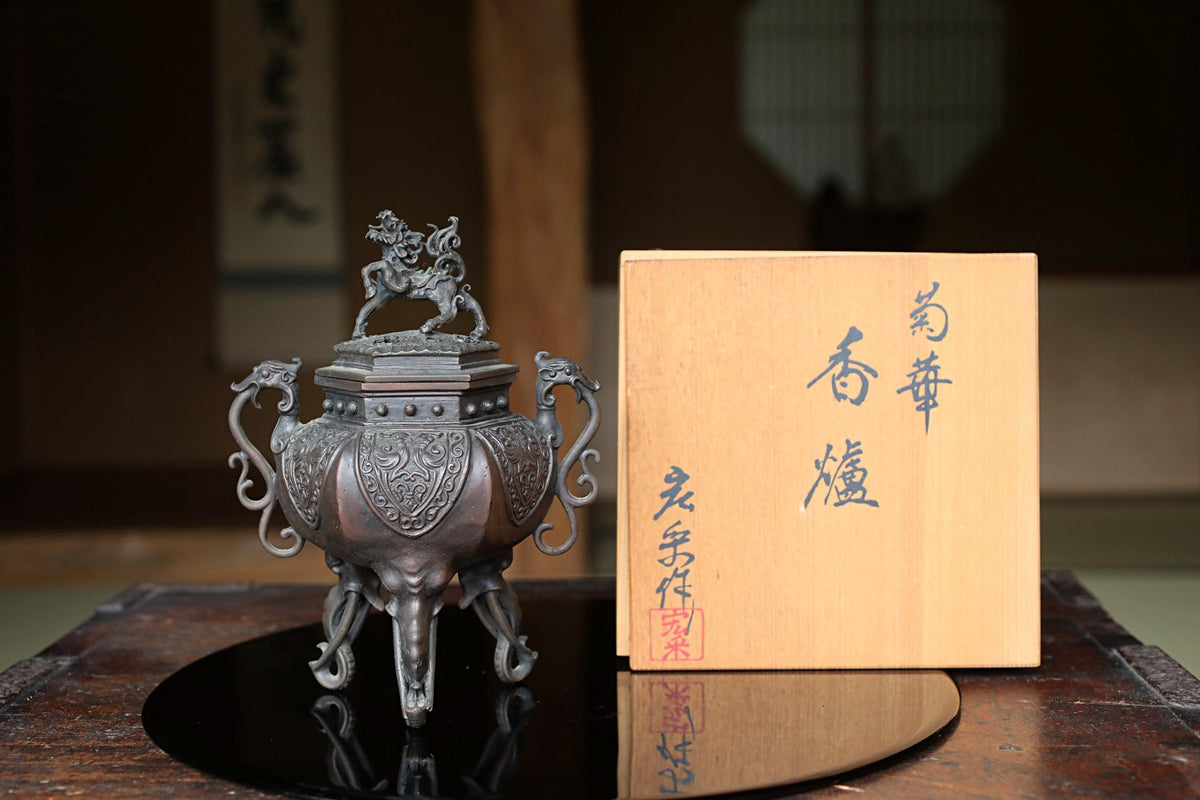 a decorative metal incense burner with an ornate lid, placed on a wooden surface next to a wooden box with Japanese characters on it.