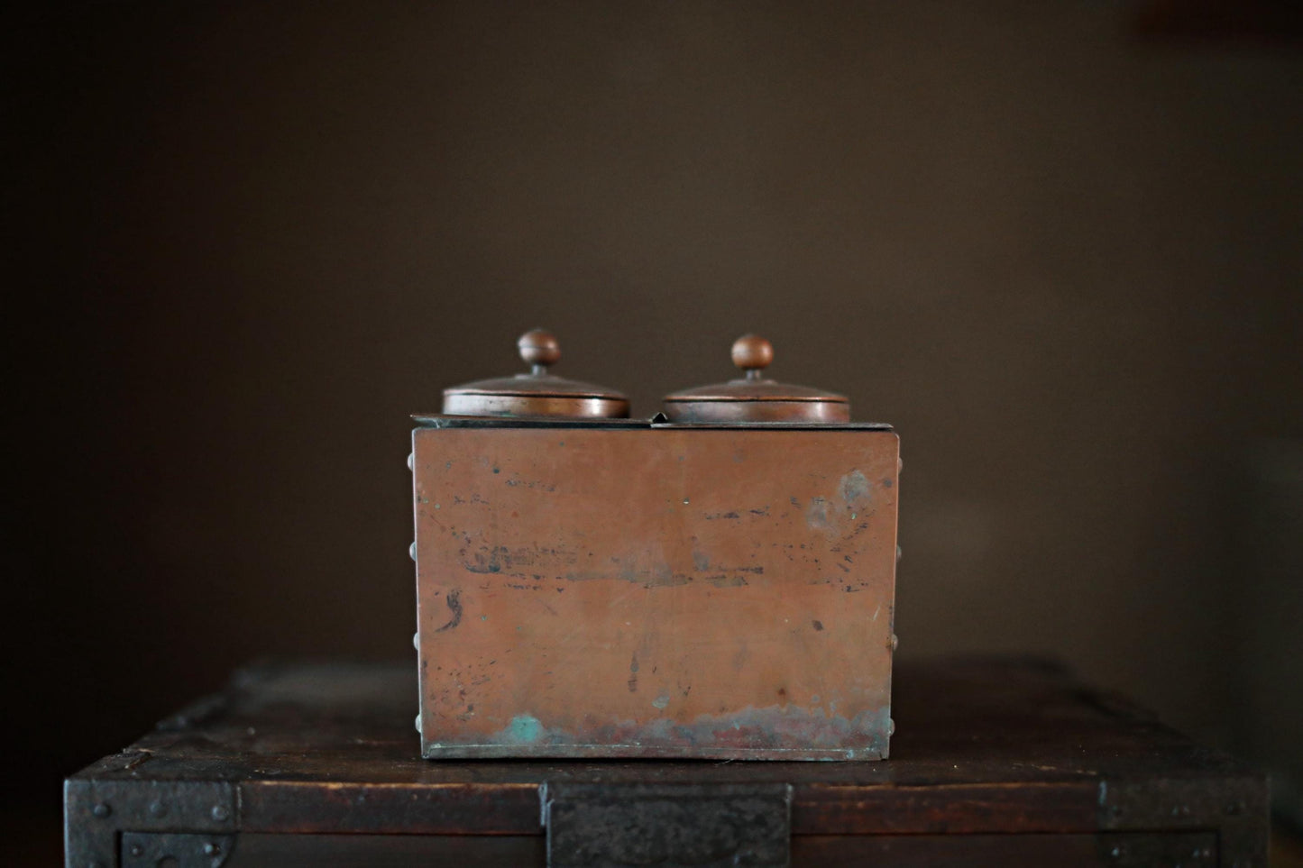 An old, weathered metal box with two domed tops sits on a wooden surface against a dark background.