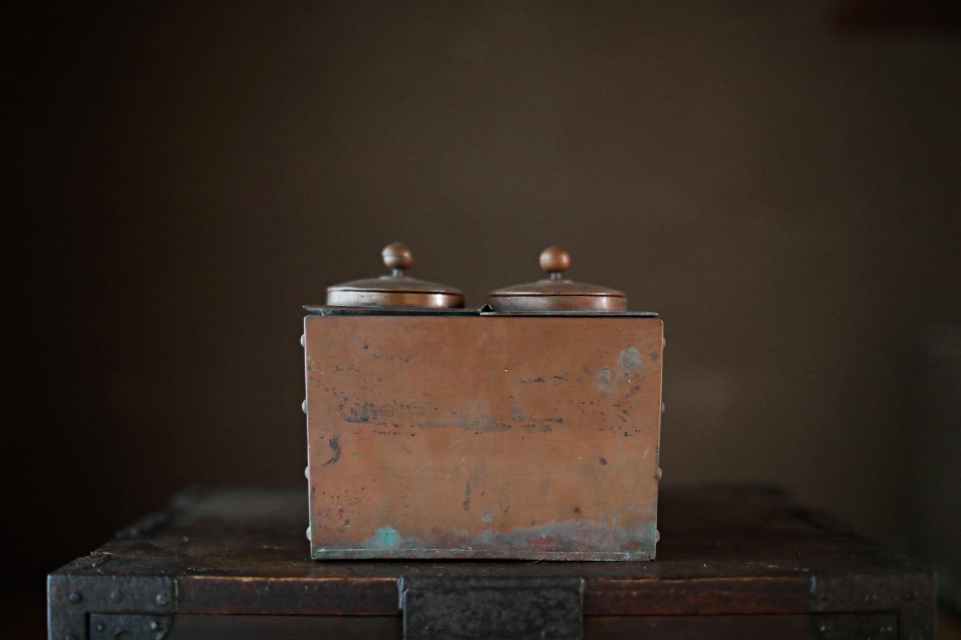 An old, weathered metal box with two domed tops sits on a wooden surface against a dark background.