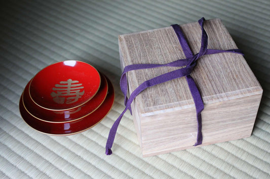a wooden box tied with a purple ribbon, next to a stack of three red plates with gold trim.