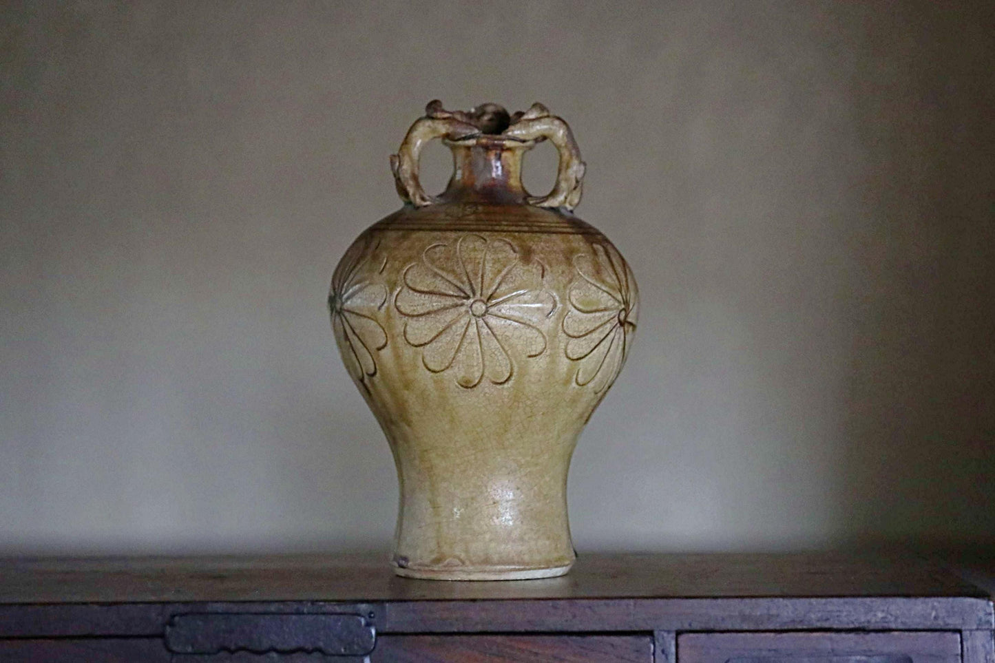 A ceramic vase with a floral design on its side, sitting on a wooden surface against a plain background.