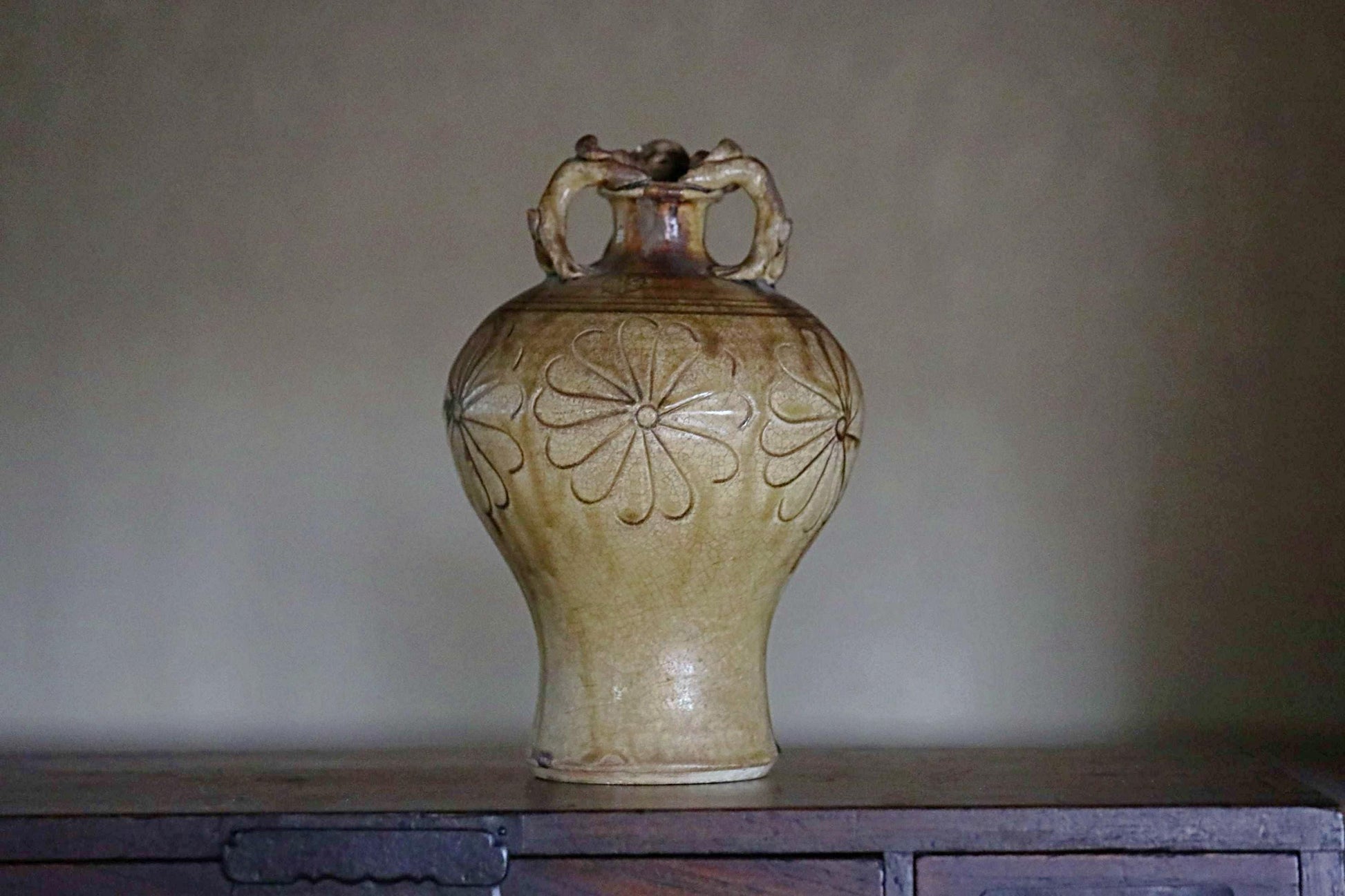 A ceramic vase with a floral design on its side, sitting on a wooden surface against a plain background.
