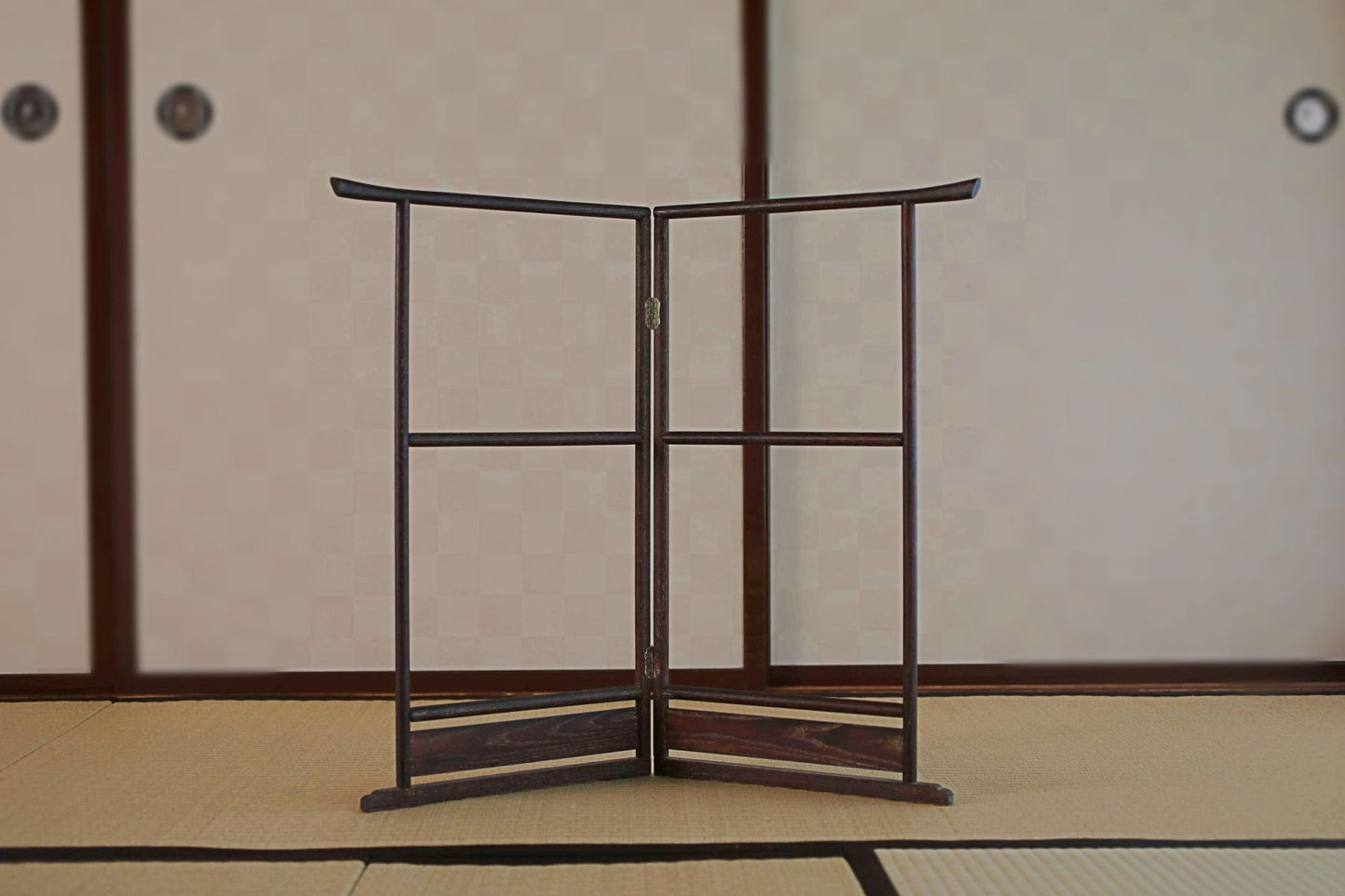 A wooden folding screen or gate with a curved top and bottom stands in the center of a room with a tatami mat floor and a traditional Japanese-style wall.