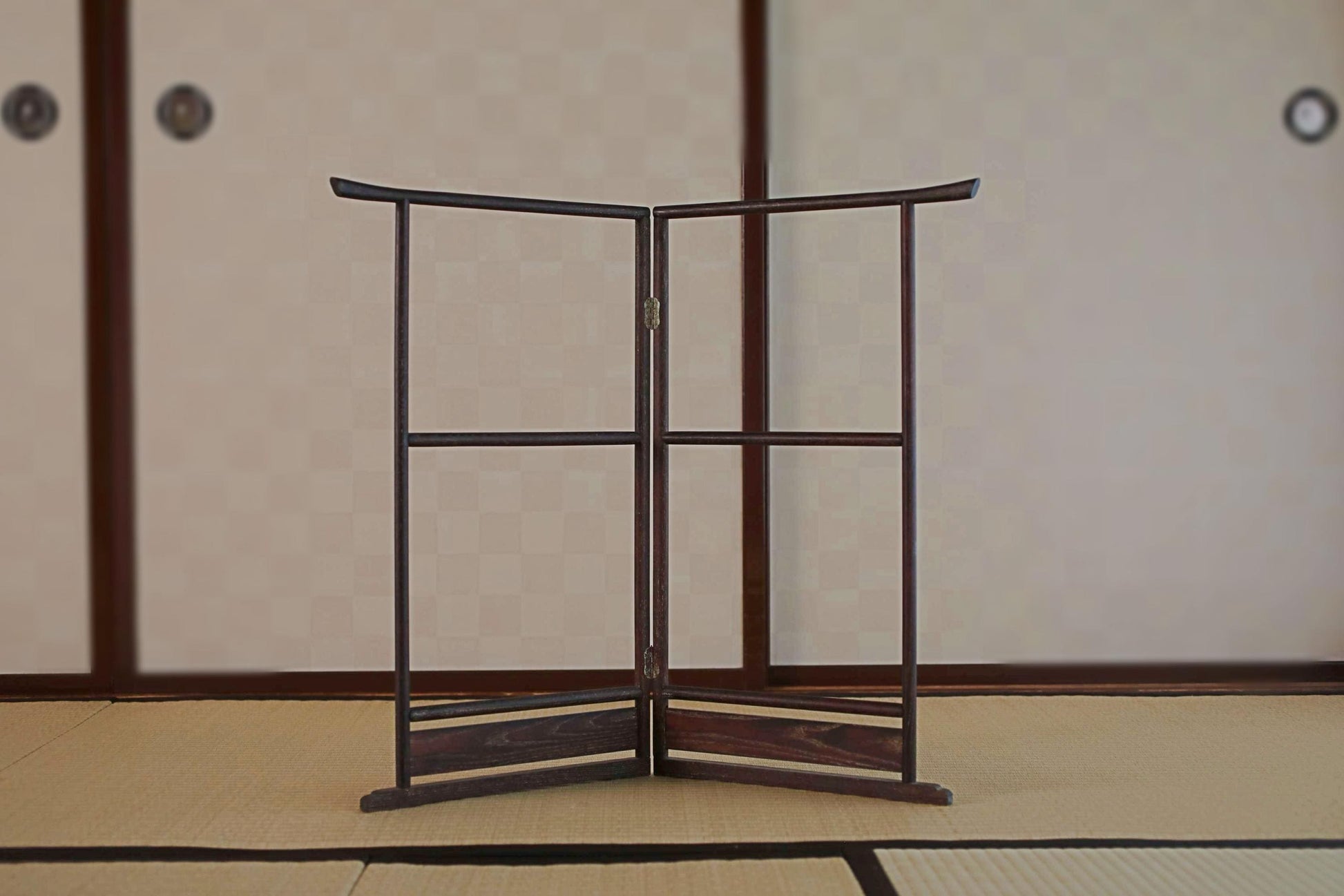 A wooden folding screen or gate with a curved top and bottom stands in the center of a room with a tatami mat floor and a traditional Japanese-style wall.