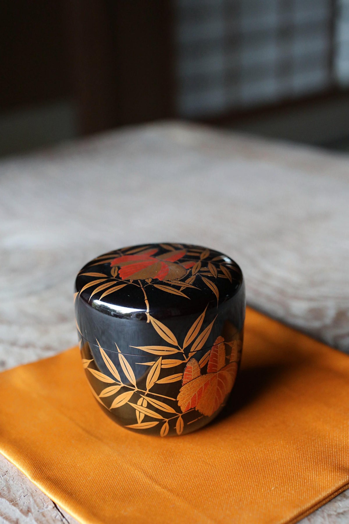 A decorative ceramic container with a black and gold floral pattern sits on an orange cloth on a wooden surface.