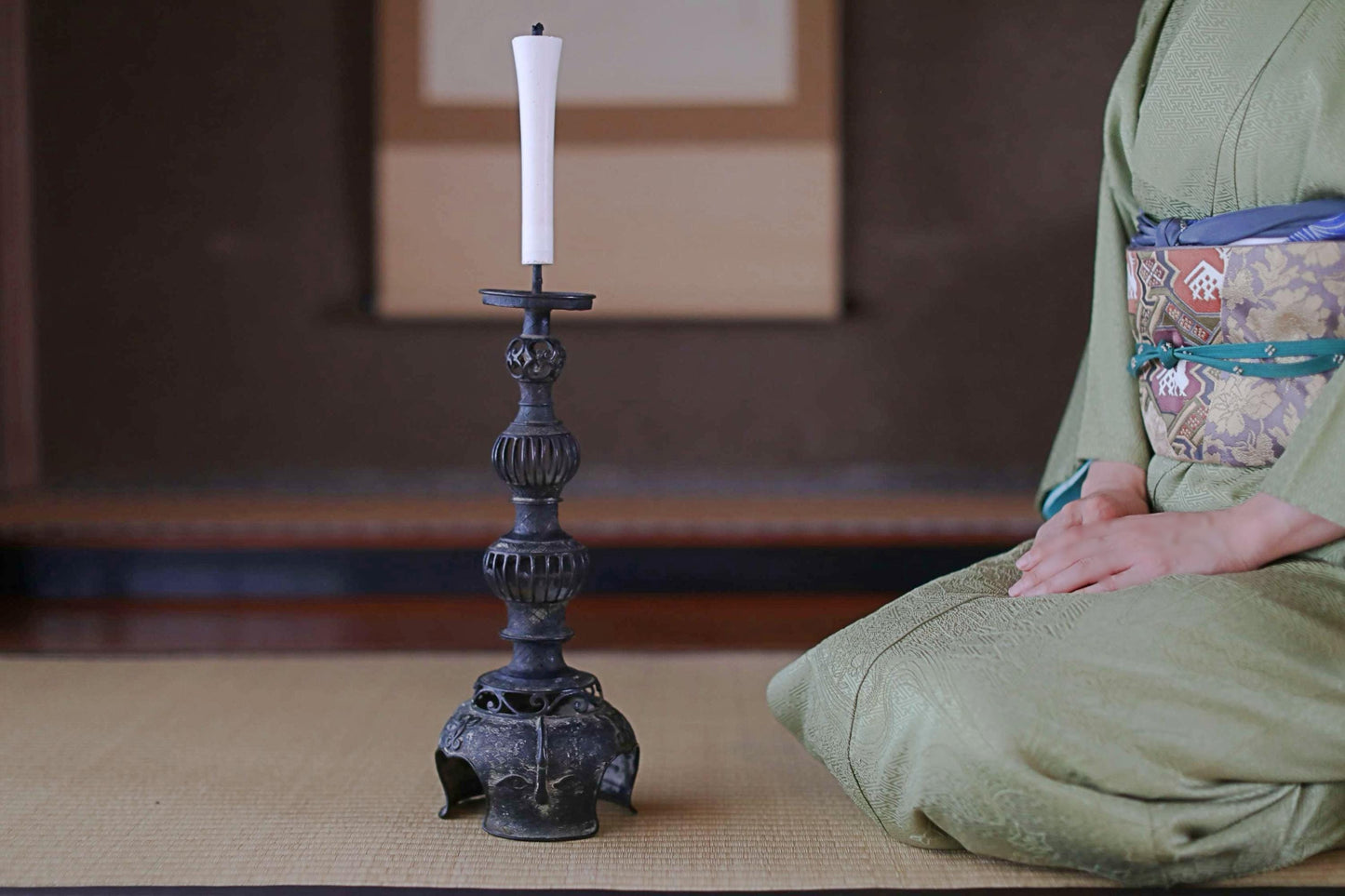 A person in a green kimono sits on a mat, with a black candlestick and a white candle standing next to them.
