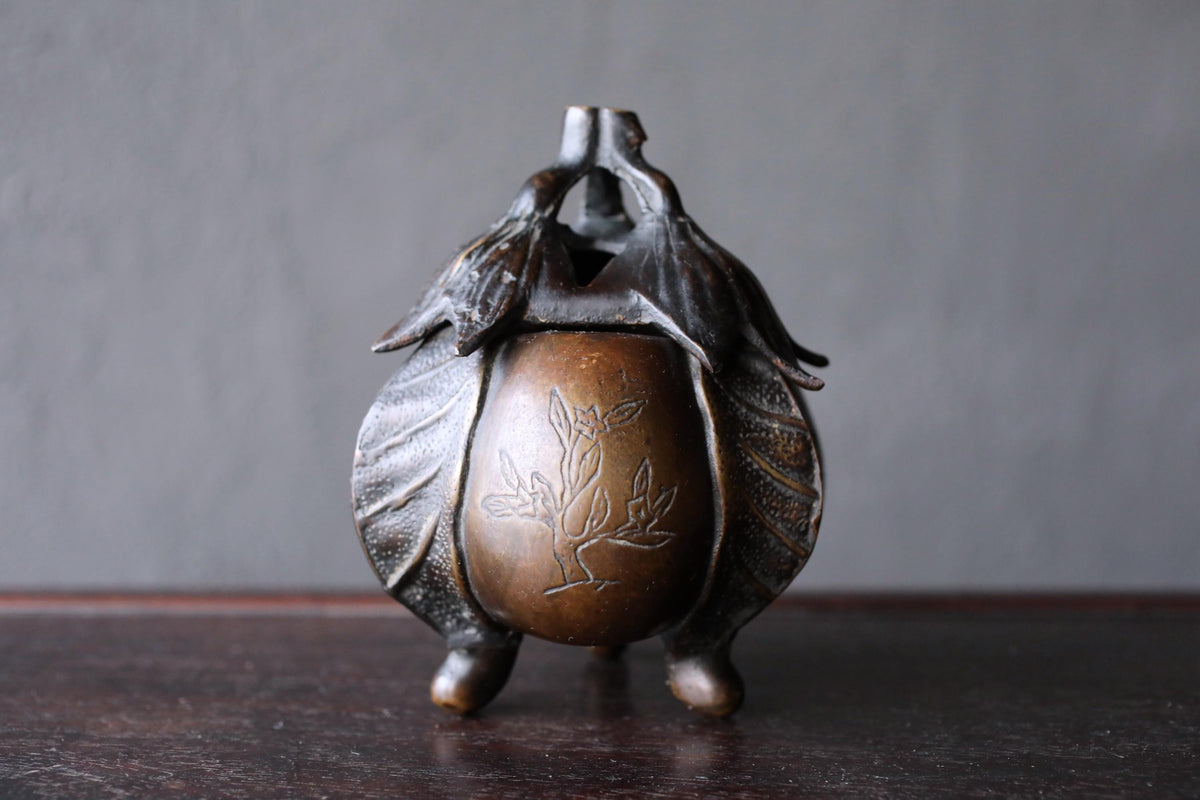 A bronze incense burner with a leaf design on the front sits on a dark wooden surface.