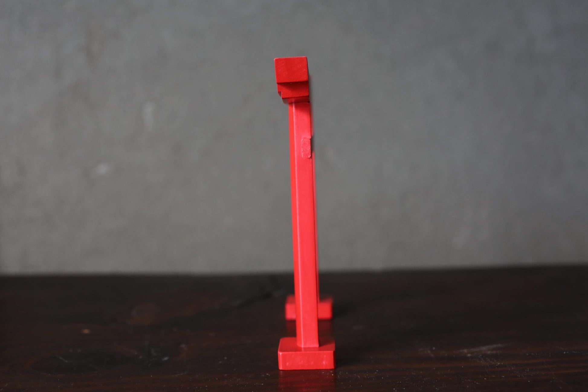 A red metal object, possibly a sculpture or a piece of furniture, is standing upright on a dark surface against a gray background.