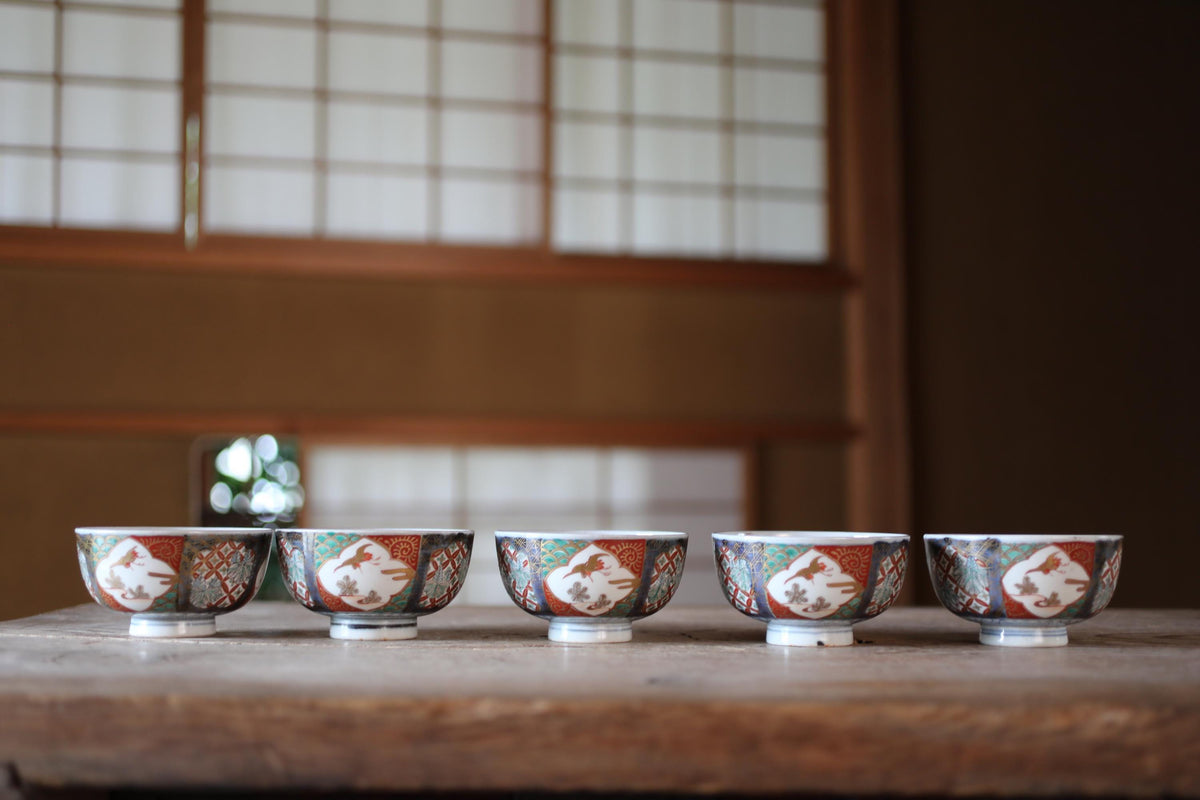 A row of five ornate bowls with intricate designs, placed on a wooden surface in front of a window with a traditional Japanese-style screen.