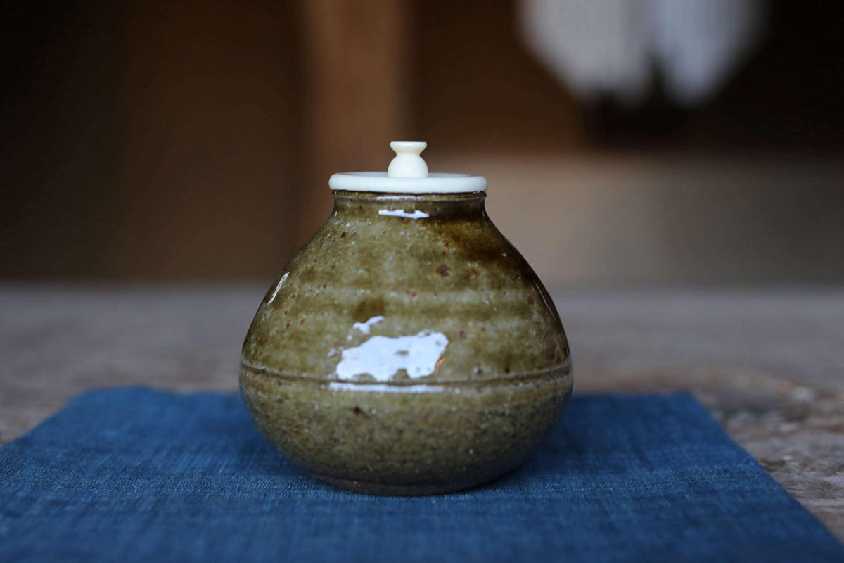 A ceramic jar with a white lid sits on a blue cloth, with a blurred background.