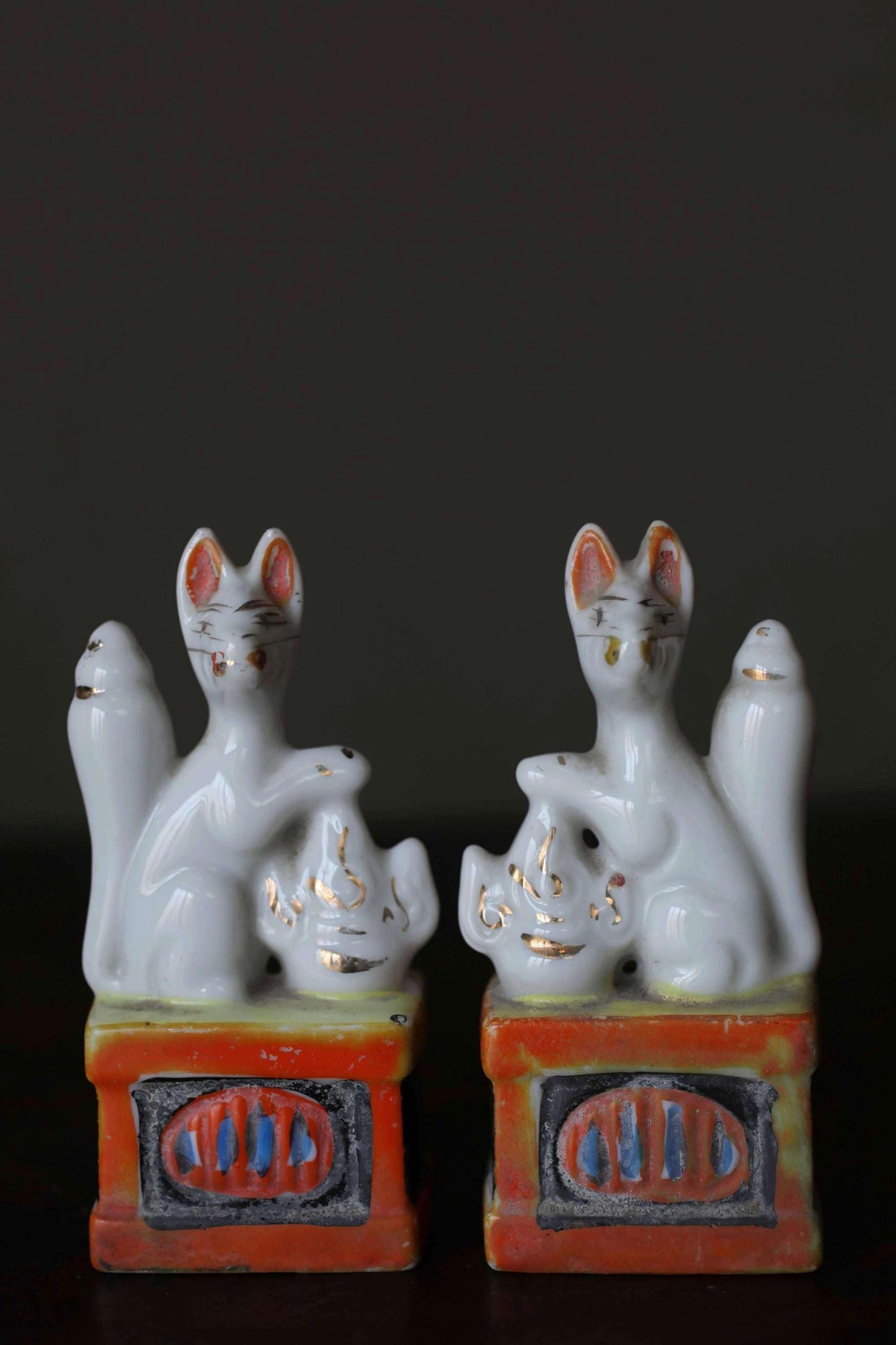 Two white ceramic figurines of cats sitting on a red and orange base, with gold accents.
