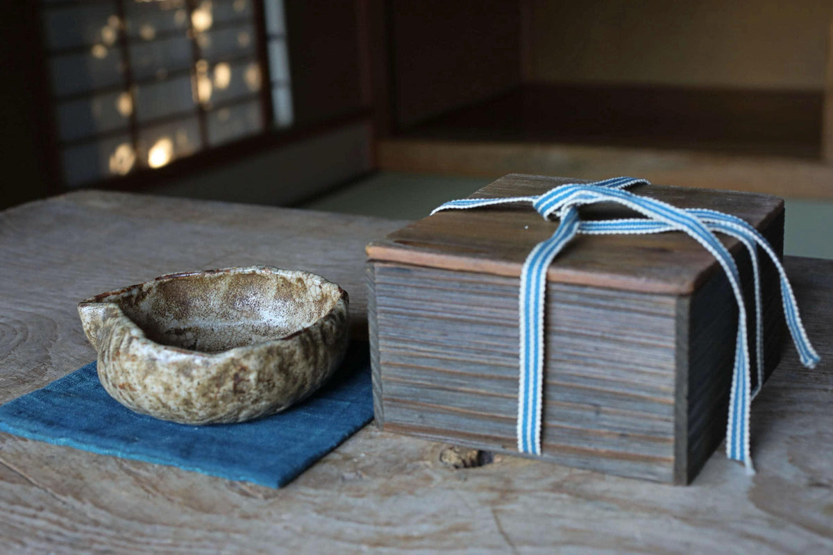a wooden box with a blue ribbon tied around it, placed on a wooden surface next to a small ceramic bowl.