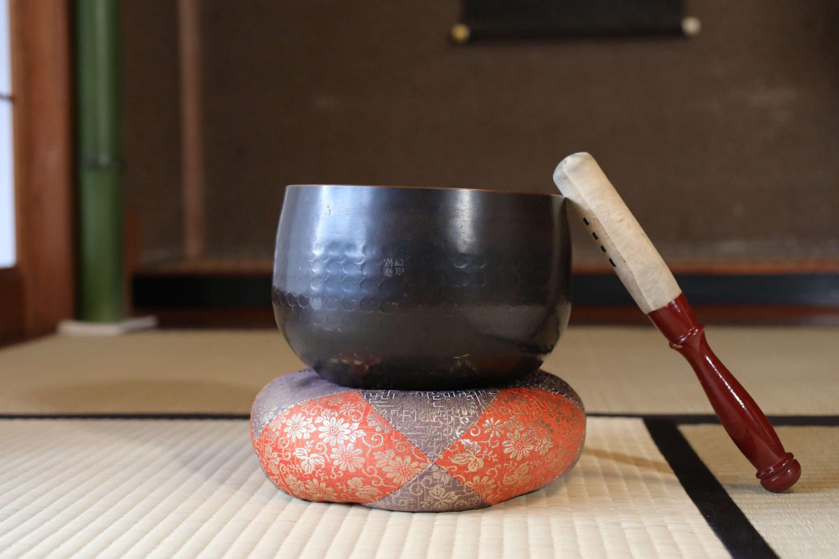 A black bowl with a wooden handle rests on a red and orange cushion, accompanied by a wooden stick, all placed on a tatami mat.