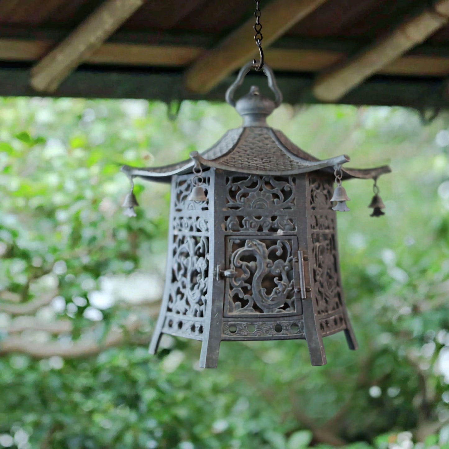 A decorative metal lantern with intricate designs hangs from a wooden structure, surrounded by lush greenery.