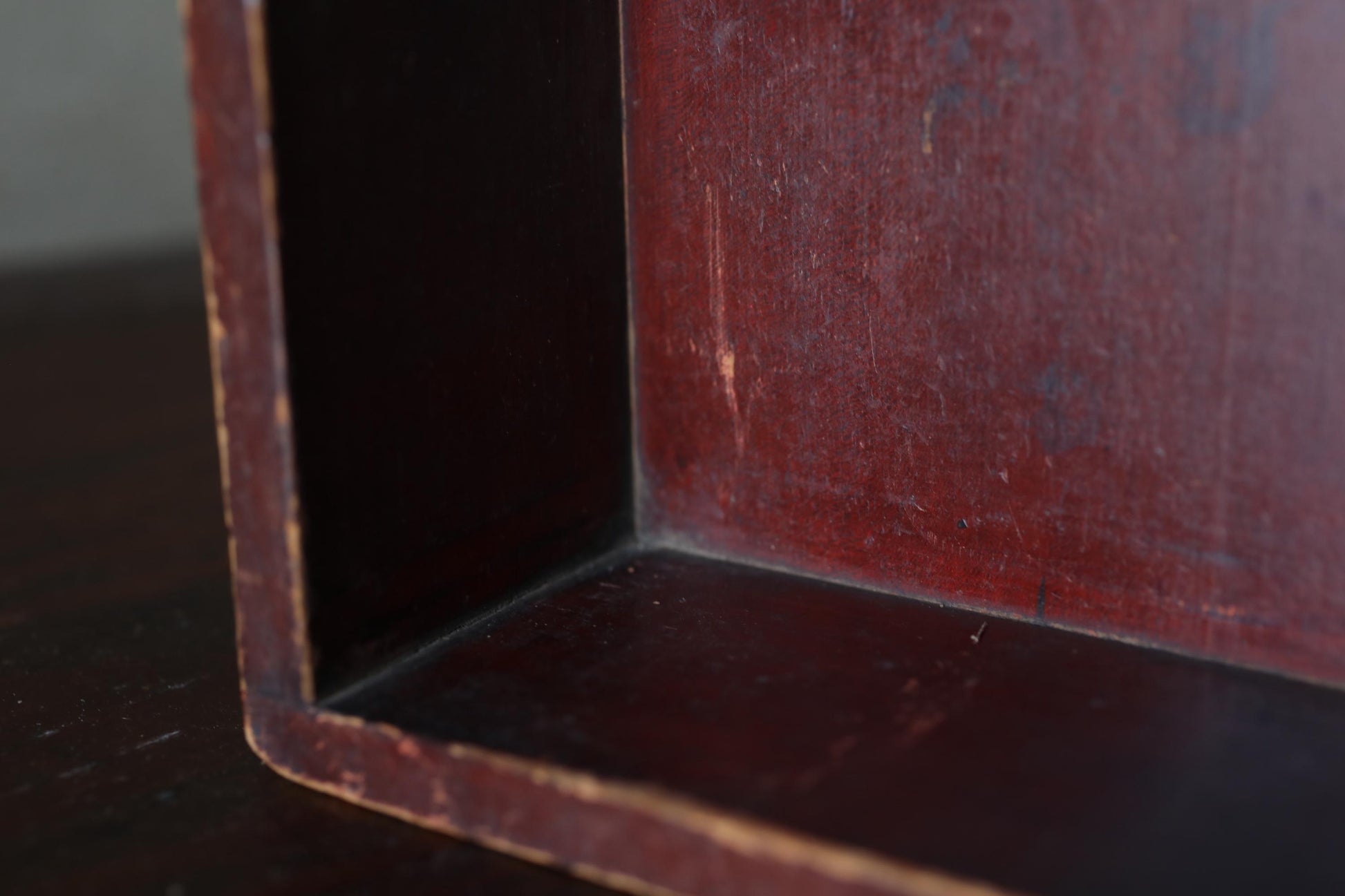 A close-up view of a dark wooden surface, possibly a shelf or cabinet, with a reddish-brown hue. The surface appears to be made of wood and has a textured, weathered appearance.