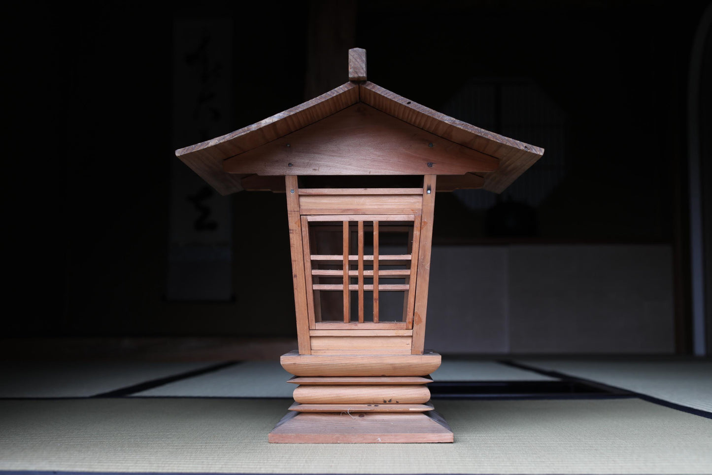 A wooden lantern with a pointed roof, featuring a lattice design, is displayed in a dimly lit room.