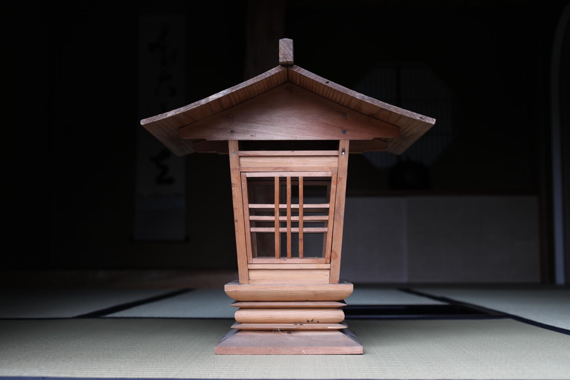A wooden lantern with a pointed roof, featuring a lattice design, is displayed in a dimly lit room.