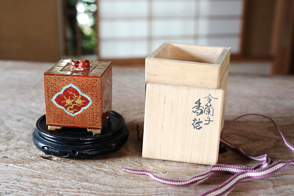 a wooden box with a red and gold design, placed on a wooden surface next to a wooden box with a lid, both of which are sitting on a black stand.