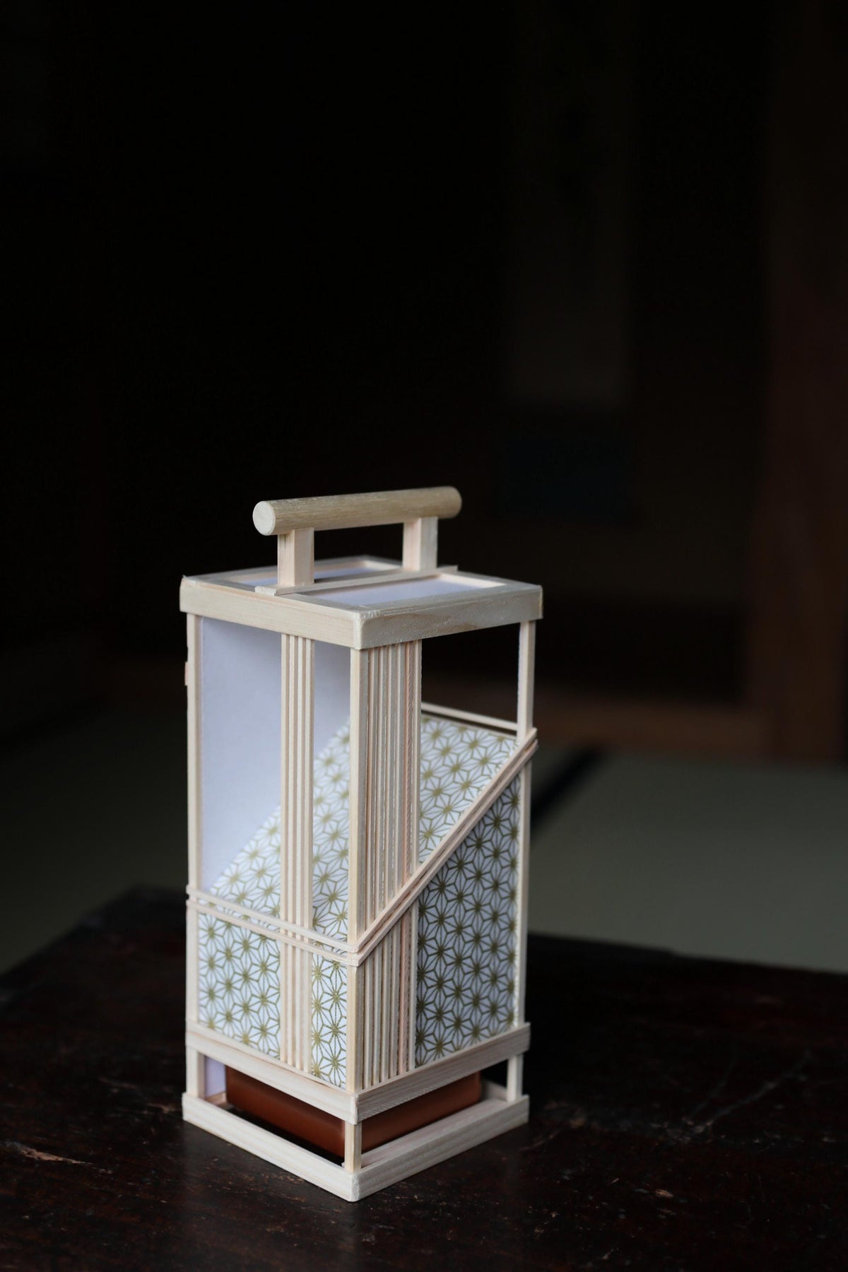 A wooden lantern with a white handle and a patterned paper covering, sitting on a dark surface.