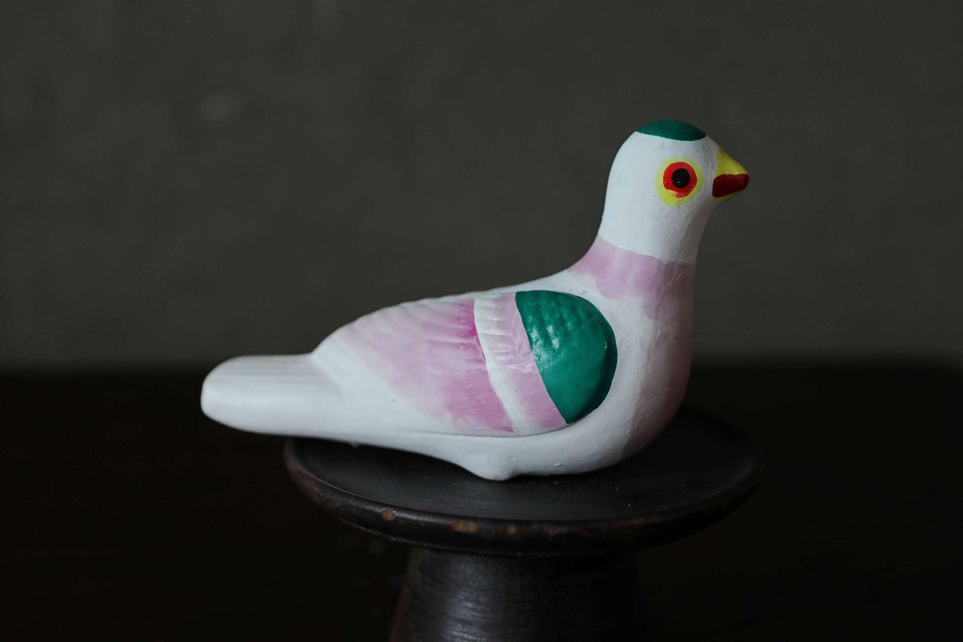 A colorful ceramic bird figurine is sitting on a black stand against a dark background.