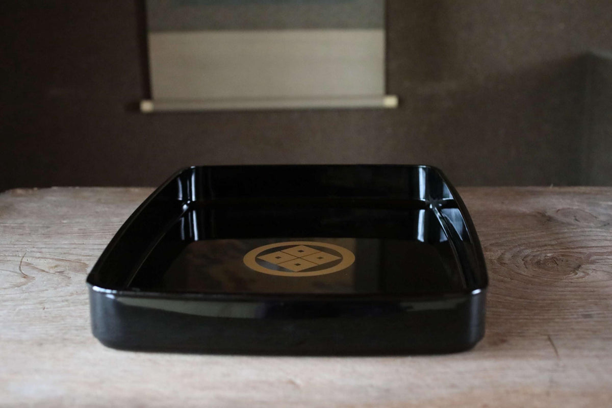 A black rectangular tray with a gold emblem on it, placed on a wooden surface.