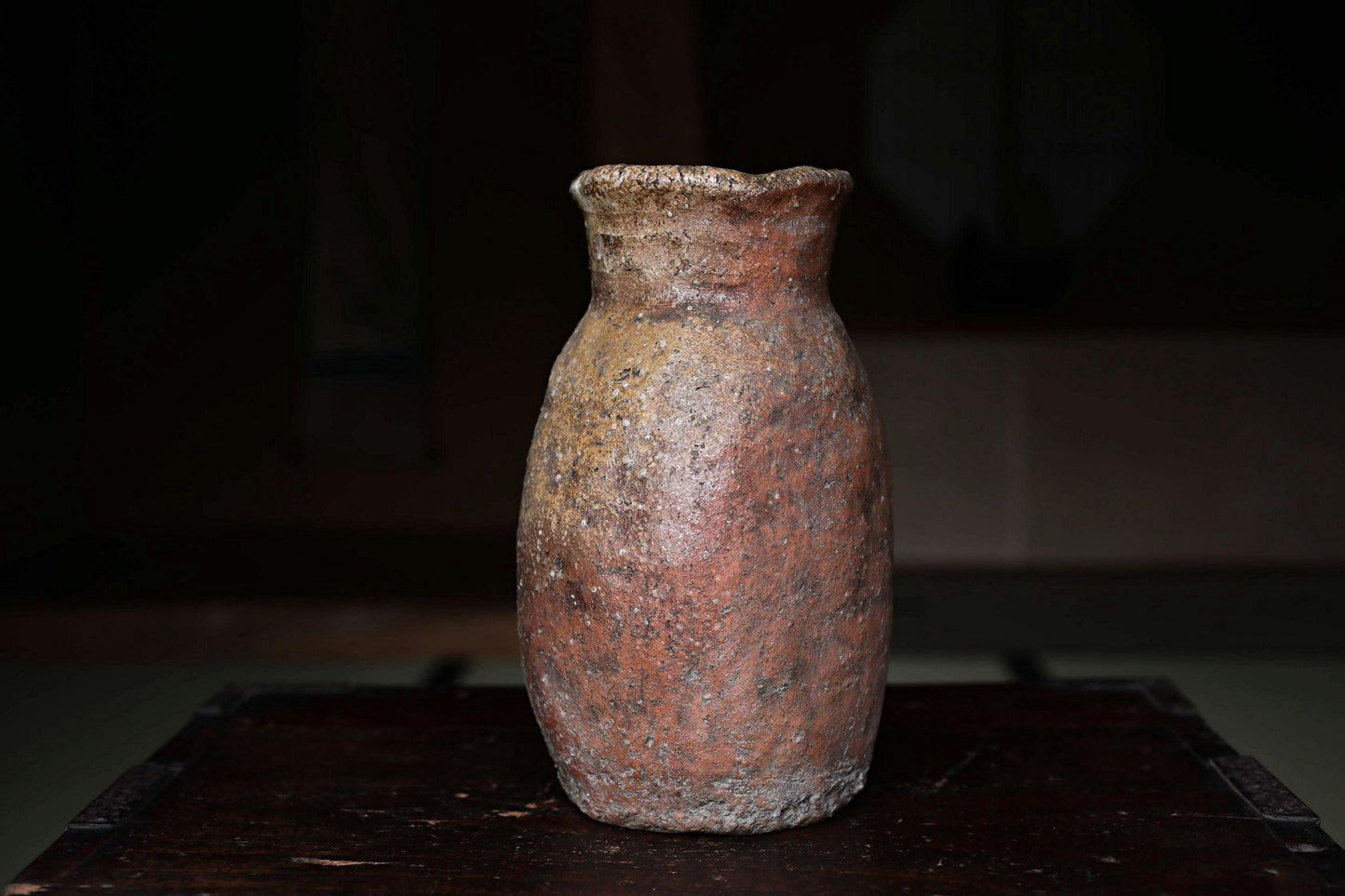 An old, weathered clay vase sits on a dark wooden surface, its reddish-brown color and rough texture suggesting it has been used for an extended period.