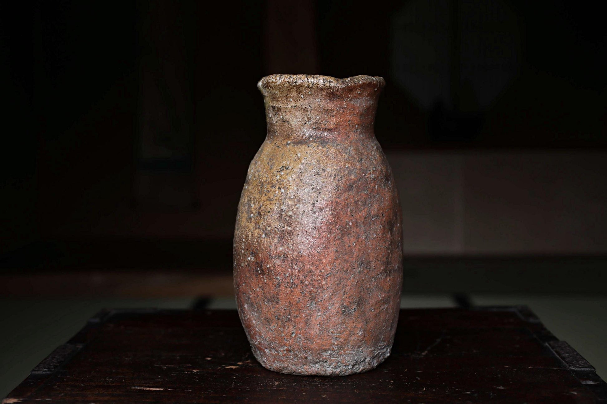 An old, weathered clay vase sits on a dark wooden surface, its reddish-brown color and rough texture suggesting it has been used for an extended period.