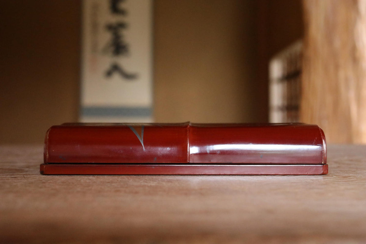 A red wooden box rests on a wooden surface, with a blurred background that includes a wall with Japanese characters.
