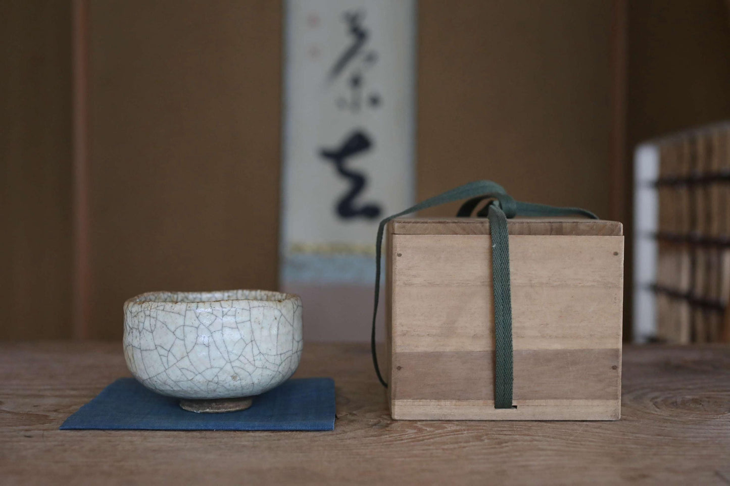 A white ceramic bowl with a green handle sits on a blue placemat, accompanied by a wooden box with a green strap on a wooden table.