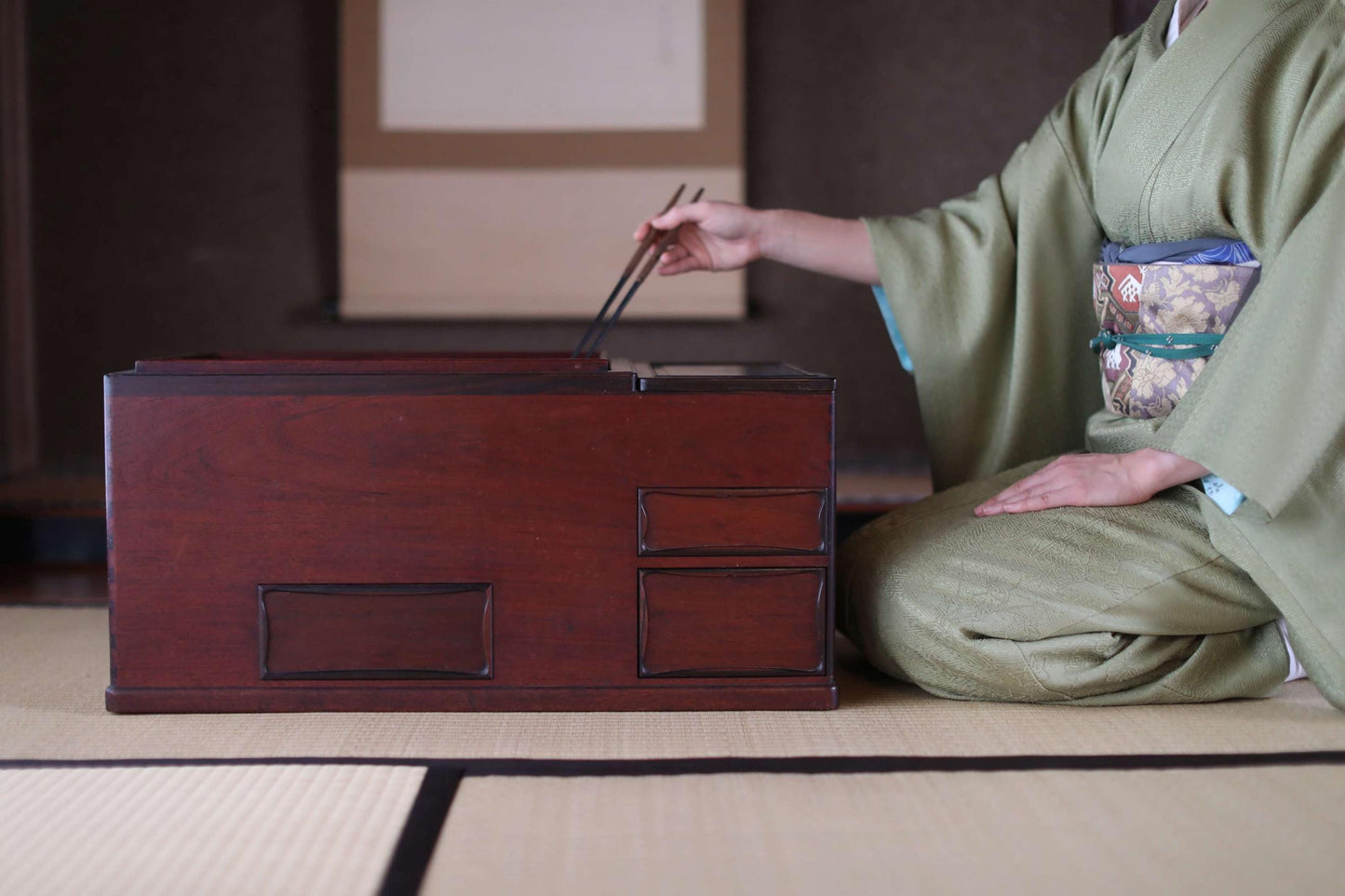 A person in a green kimono is kneeling on a tatami mat, using chopsticks to pick up something from a wooden box.