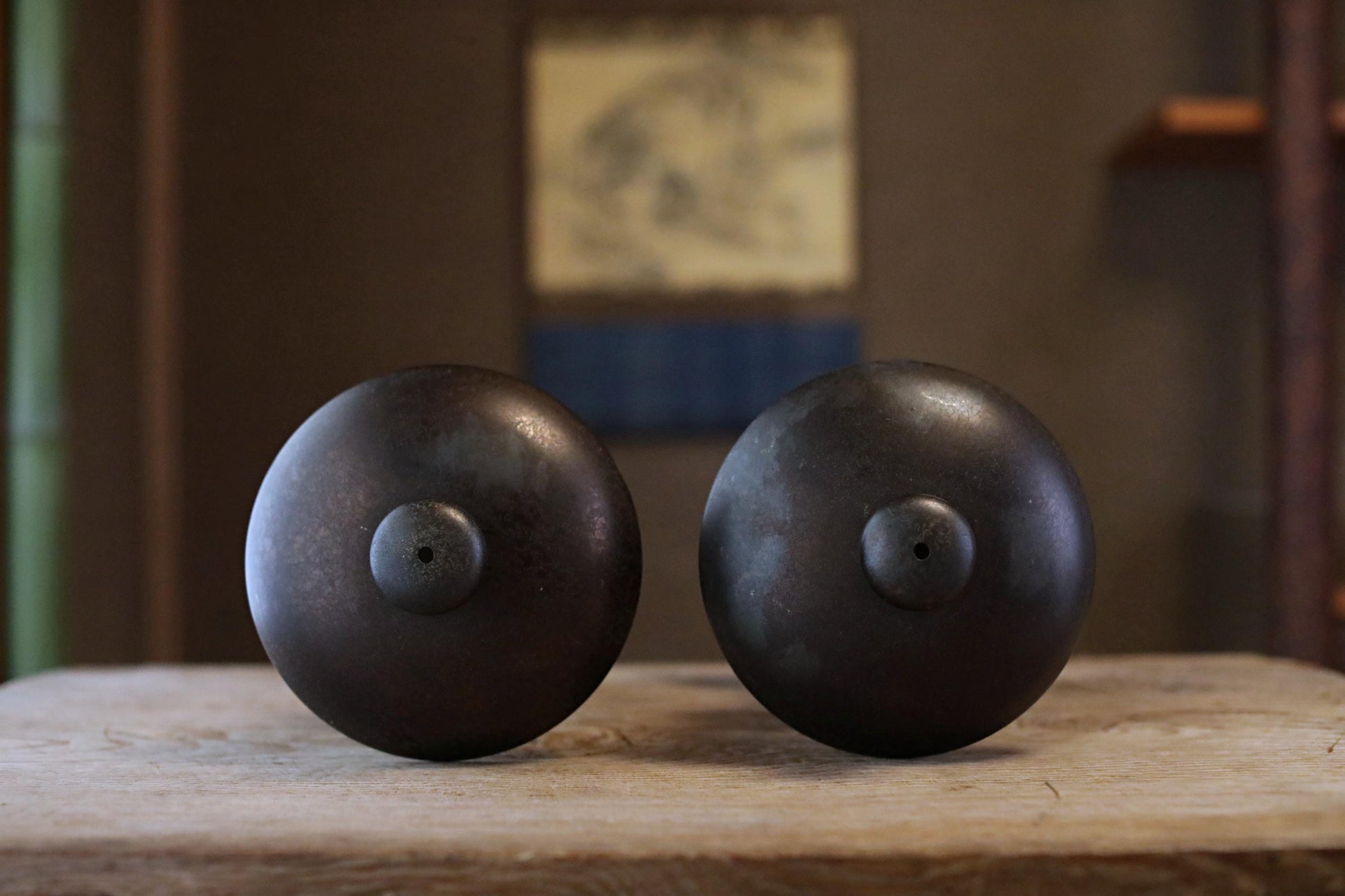 Two black metal objects with round tops are placed on a wooden surface, with a blurred background that includes a painting.
