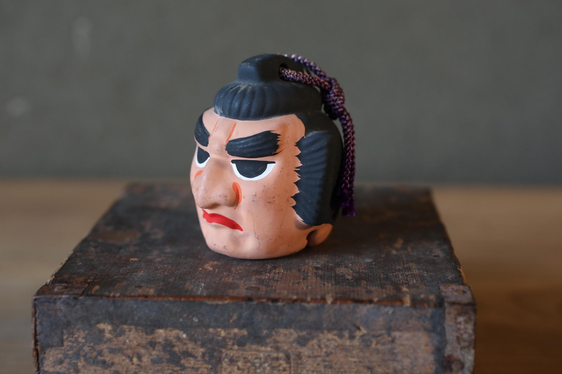 A ceramic figurine of a man's head with a black hairpiece and a red mouth, sitting on a wooden surface.