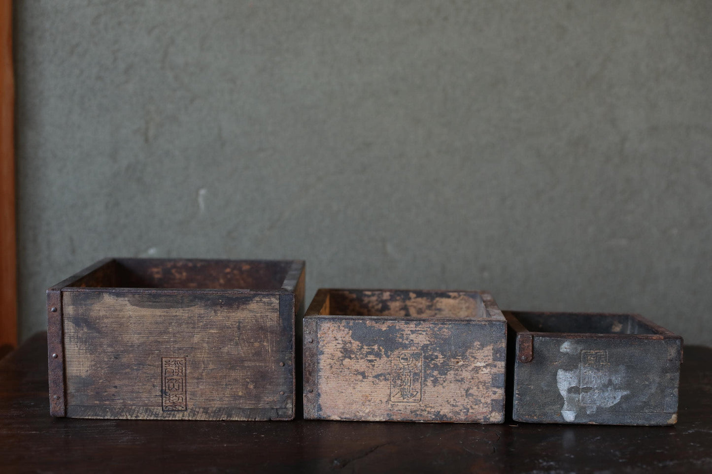 Three old, weathered wooden crates of varying sizes sit on a dark surface against a gray wall.