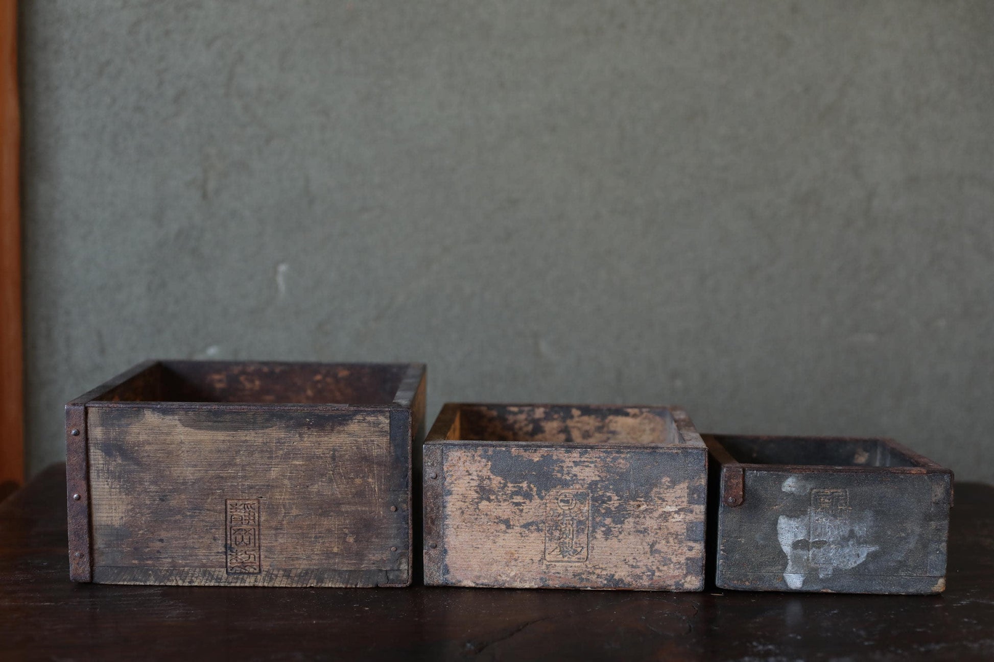 Three old, weathered wooden crates of varying sizes sit on a dark surface against a gray wall.