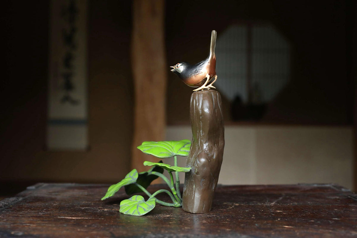 A small bird figurine perched on a brown wooden post, with green leaves sprouting from the base, sits on a wooden surface.