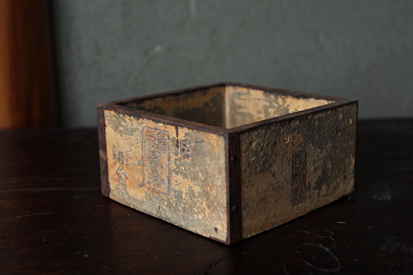 An old, weathered wooden box sits on a dark surface, with a textured and worn appearance.