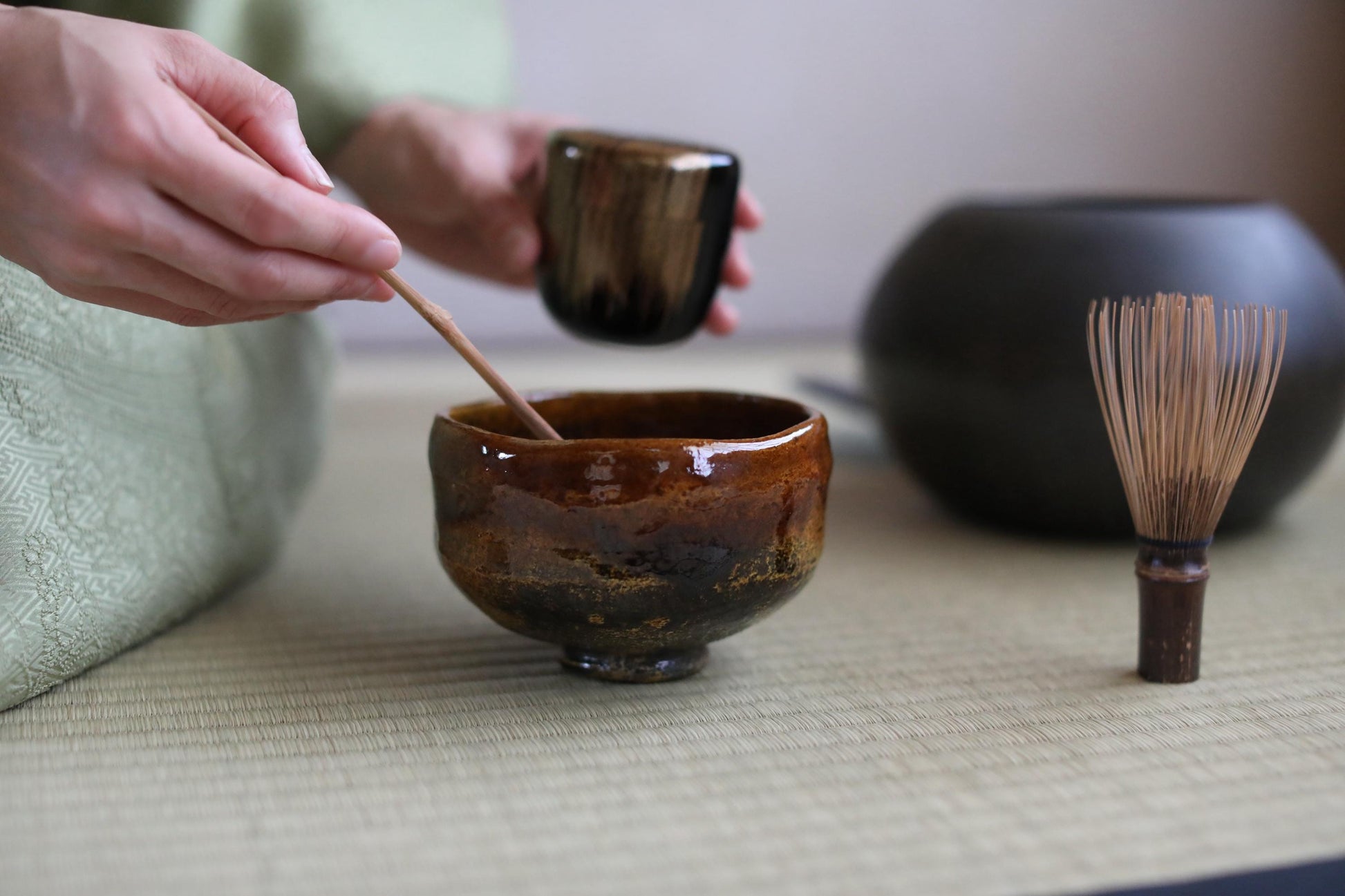 A person's hands are shown stirring a brown ceramic bowl with a wooden spoon, while a black ceramic bowl and a small wooden whisk are visible in the background.