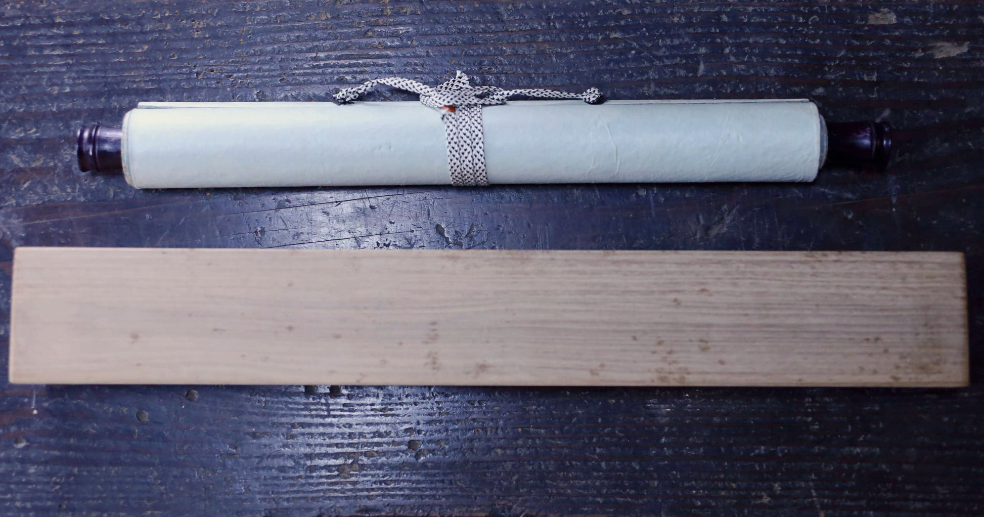 A scroll of parchment paper and a wooden plank are placed on a dark wooden surface.