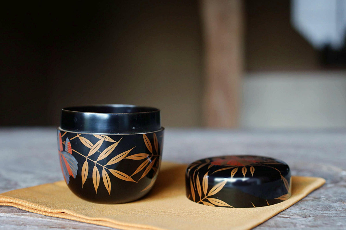 A black ceramic cup with a leaf design sits on a yellow cloth, accompanied by a lid with a similar leaf design.