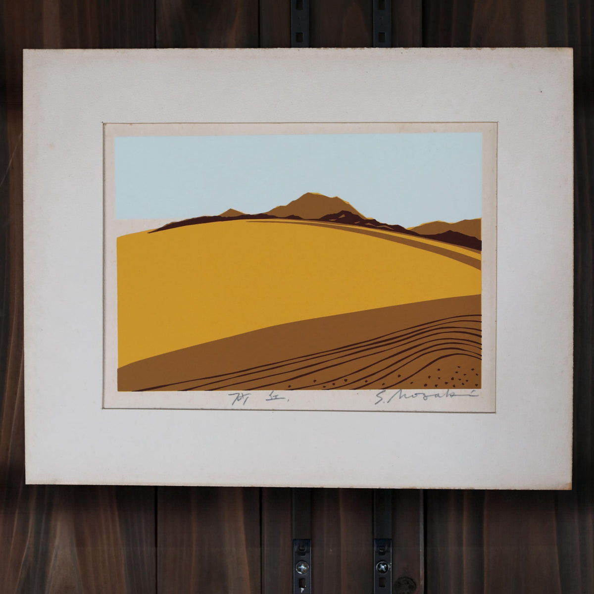 The image is a framed artwork depicting a landscape with a hill and mountains in the background, set against a blue sky.
