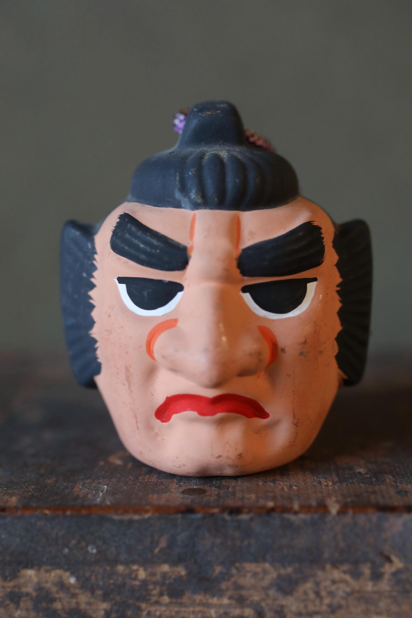 A close-up image of a toy or figurine with a face, featuring a red mouth and black hair, sitting on a wooden surface.