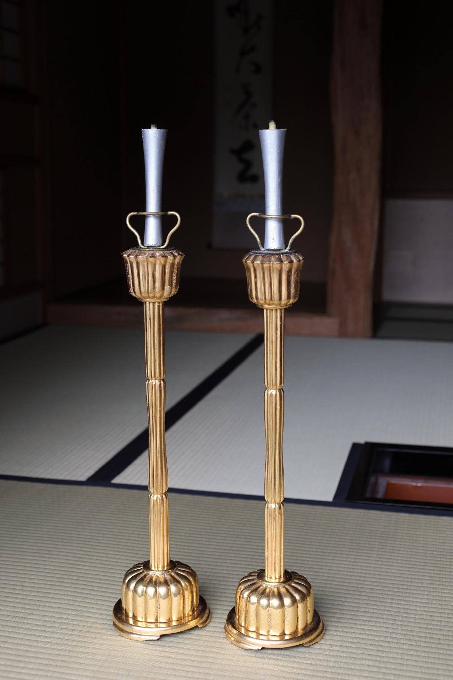 Two ornate gold candlesticks with white candles stand on a tatami mat in a dimly lit room.