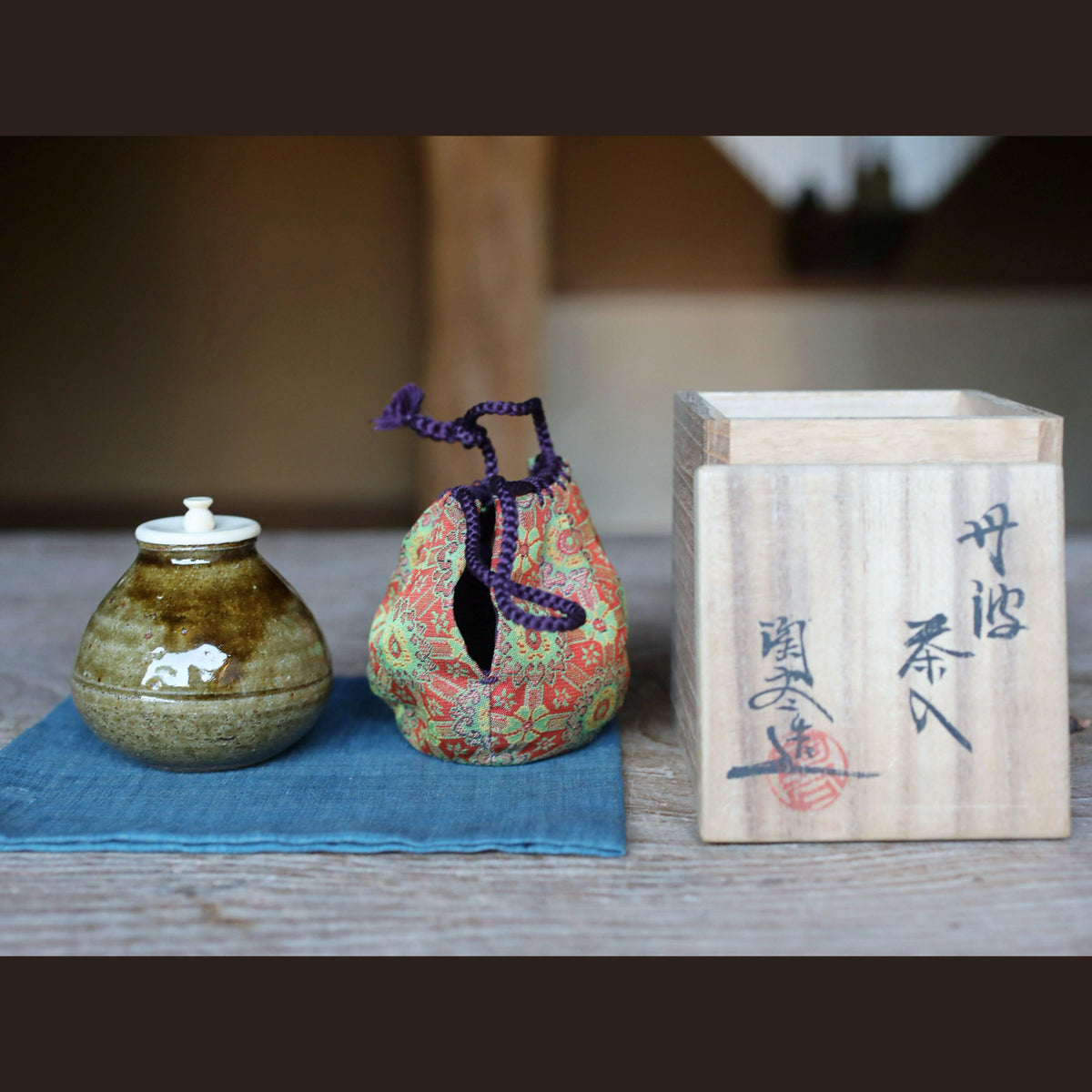 a wooden box with Japanese characters on it, placed next to a ceramic jar and a small bag with a purple string.
