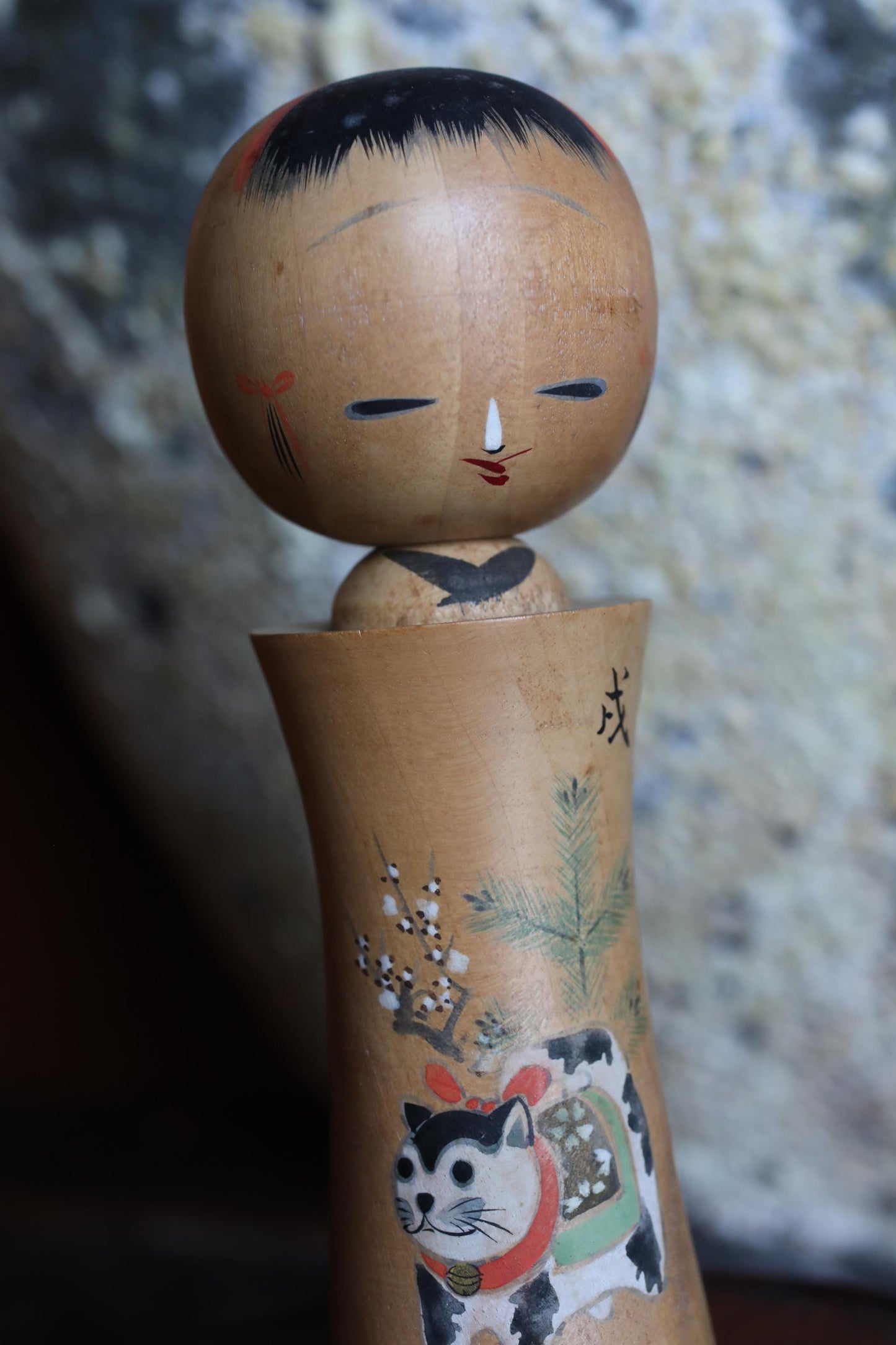 A wooden doll with a face and a cat on its body, set against a blurred background.