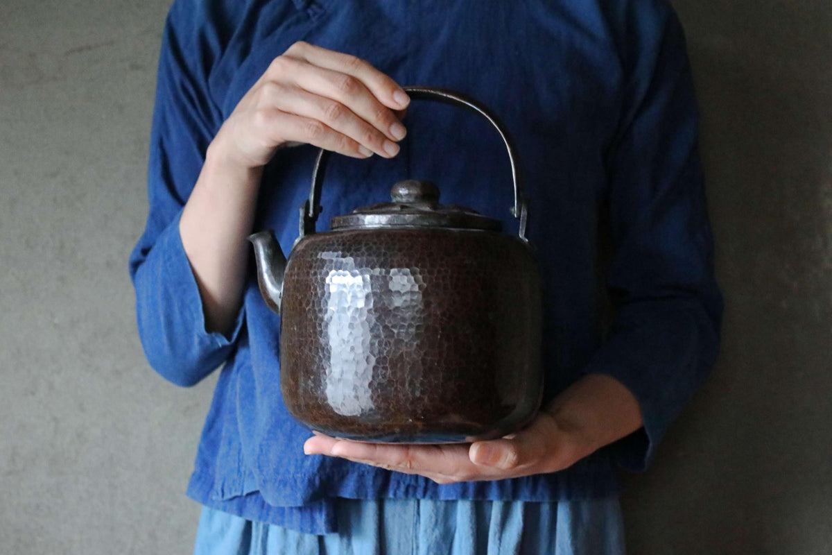A person wearing a blue shirt is holding a dark brown, textured kettle in their hands.